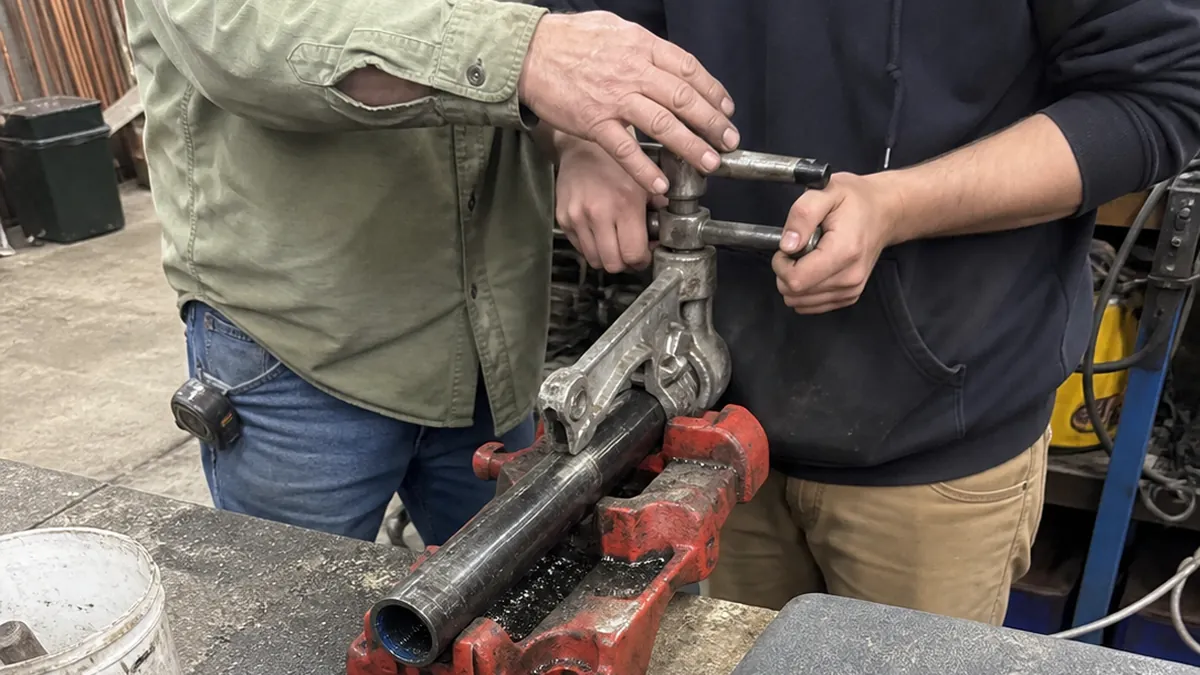 An experienced plumber and a younger helper working together at a pipe vise in a shop