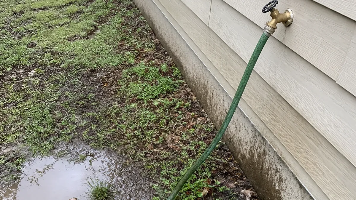 An outdoor hose bibb with a garden hose attached and the end of the hose lying in a puddle in the yard