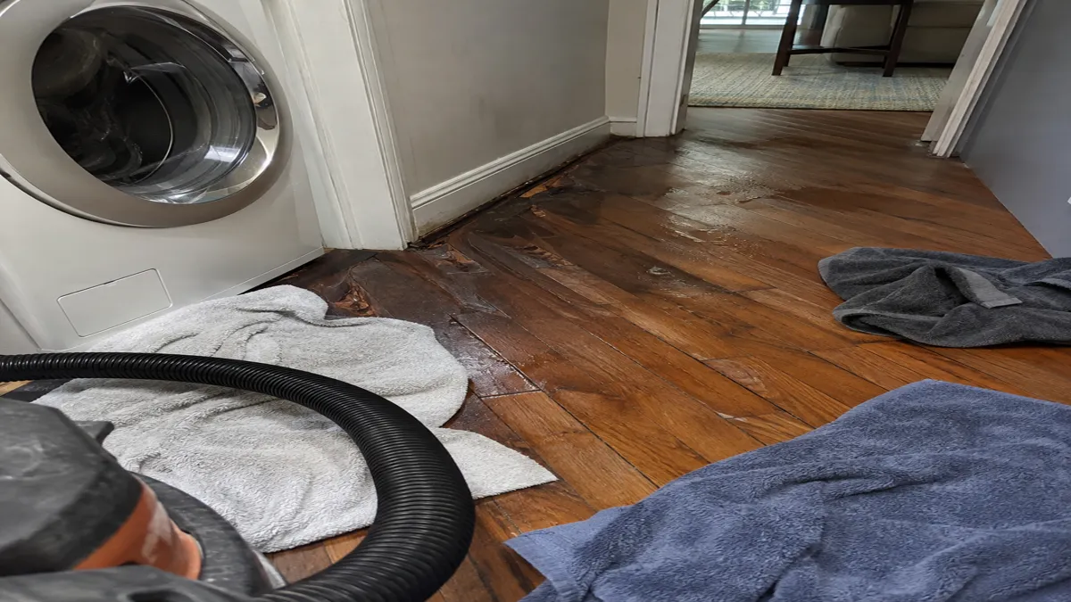 Water-damaged hardwood flooring near a washing machine, with towels and a shop-vac hose on the floor