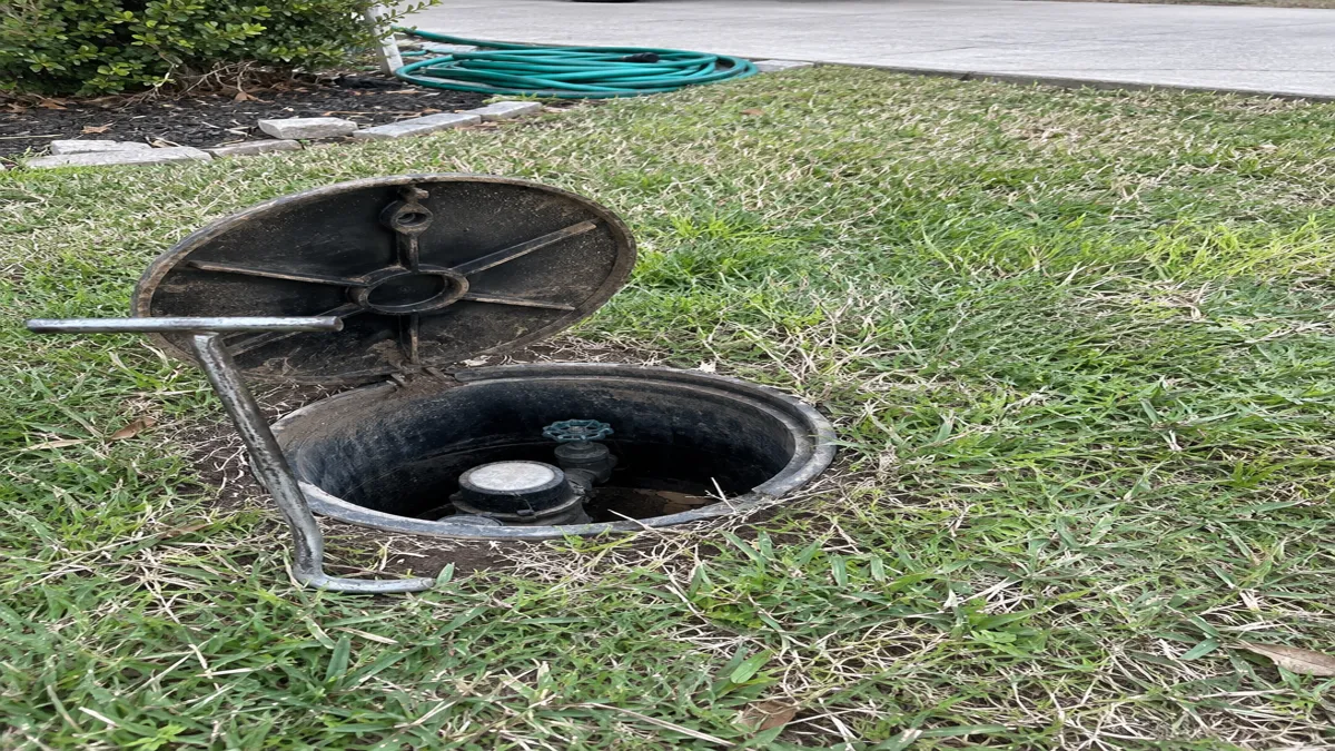 A water meter box lid lifted open in a front yard with a meter key leaning against the rim, revealing the meter and shutoff valve inside