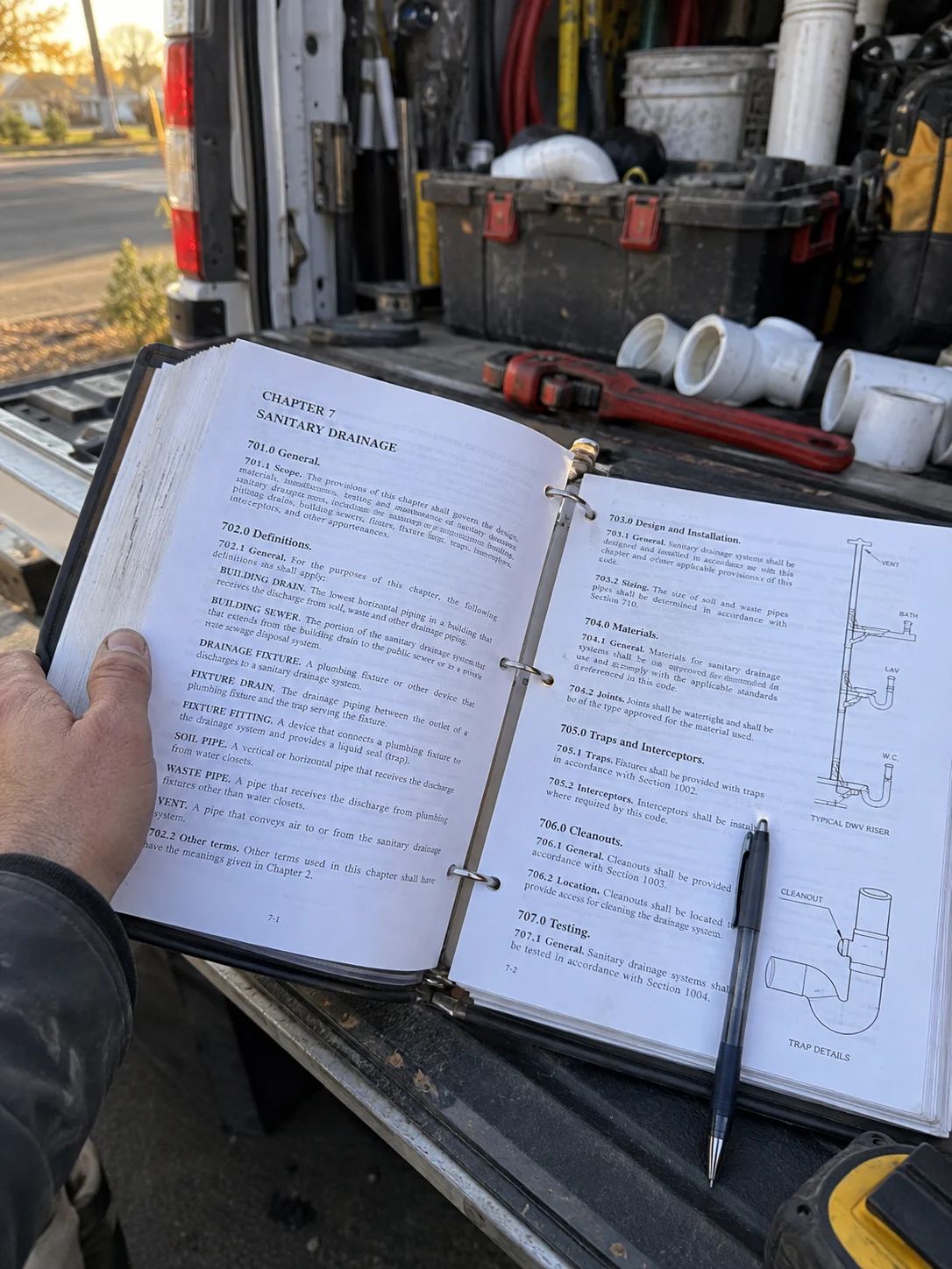 A plumber sitting on the open tailgate of his work van at the end of the day, reading a thick reference binder