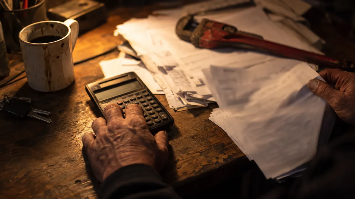 Weathered hands resting on a pocket calculator at a cluttered wooden desk piled with paperwork, lit by a single lamp, beside a coffee mug, van keys, and a red pipe wrench