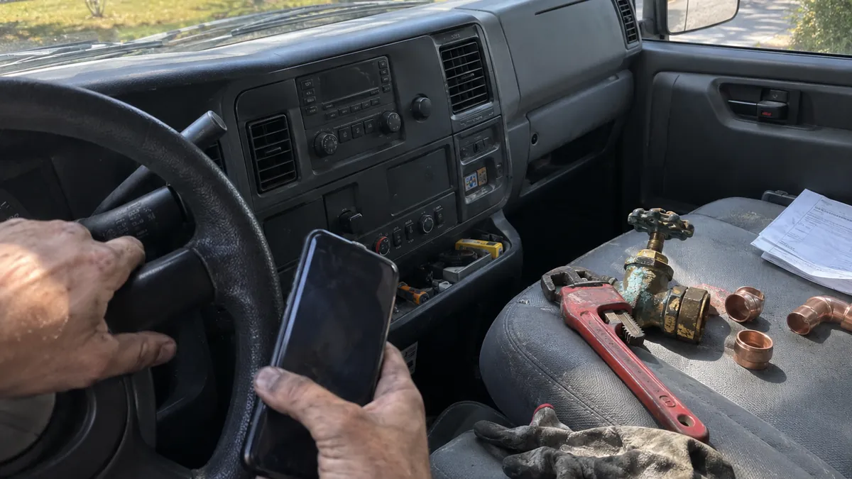 A plumber's hands in a service van cab, one on the wheel and one holding a phone, with a pipe wrench, an old brass valve, copper fittings, and work gloves on the passenger seat