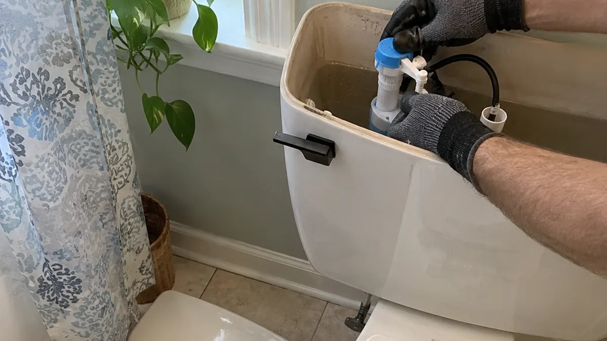 Gloved hands adjusting a new fill valve inside an open toilet tank in a home bathroom