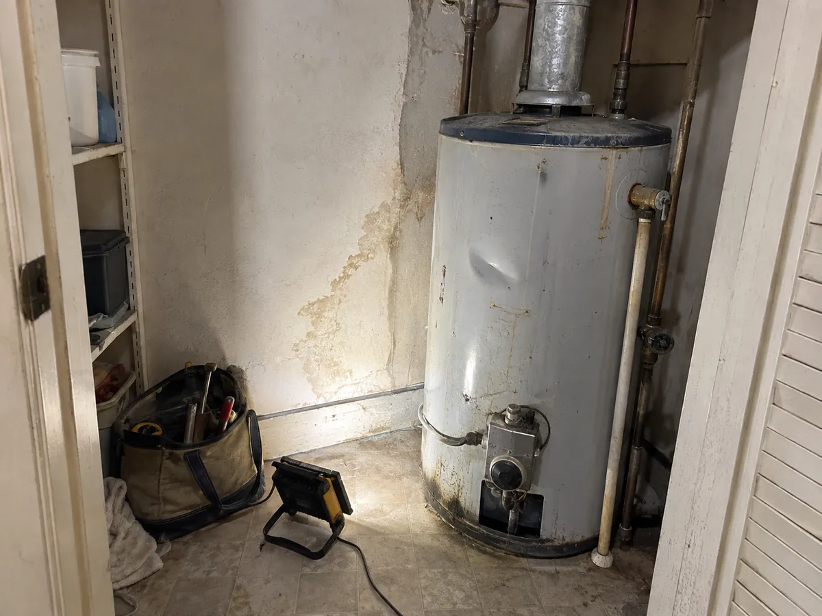A plumber's work light and tool bag on the floor of a rental unit's utility closet next to an older gas water heater