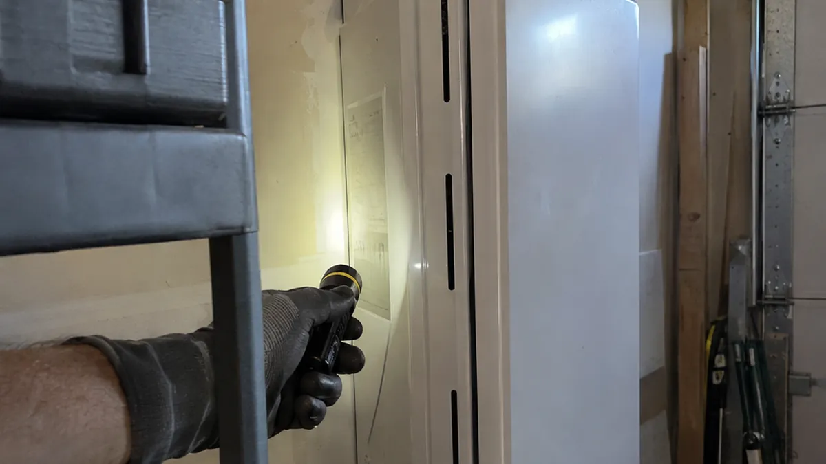 A gloved hand shining a flashlight on the side label of a white wall-mounted tankless water heater in a garage